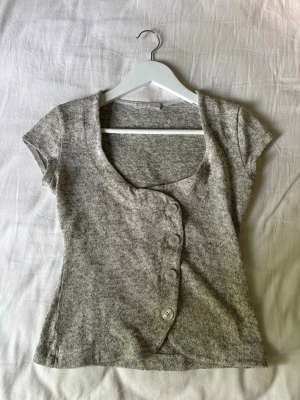 Grå kortärmad topp med knappar - Säljer en grå kortärmad topp med rundad hals och fyra stora knappar framtill. Toppen har en asymmetrisk knappslå och är tillverkad i mjukt stickat material. Perfekt för dig som gillar en enkel men unik stil.