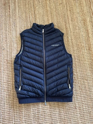 Svart dunväst från Armani Exchange - Snygg svart dunväst från Armani Exchange med diskret logga på bröstet. Västen har dragkedja framtill, två fickor med dragkedja och en hög krage. Quiltad design med diagonala sömmar ger en modern look. Perfekt för lager-på-lager och streetstyle.