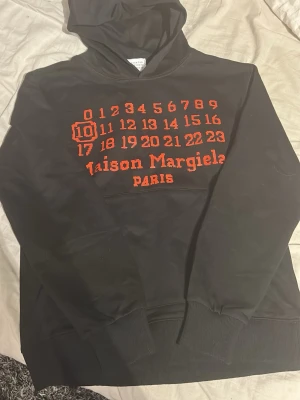Maison Margiela svart hoodie - Svart hoodie från Maison Margiela med röd text och siffror på bröstet samt logga och Paris-tryck. Klassisk huva och ribbade muddar vid ärmslut och nederkant. Tillverkad i mjukt bomullsmaterial, perfekt för en avslappnad och trendig look.