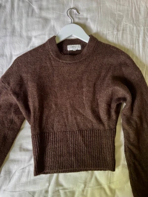 Brun stickad tröja med ribbad midja - Mysig brun stickad tröja med rund halsringning och långa ärmar. Tröjan har en bred ribbad kant vid midjan som ger en snygg siluett. Perfekt för lager på lager och enkel att matcha med jeans eller kjol.