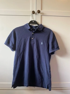Marinblå piké från Polo Ralph Lauren - Klassisk marinblå pikétröja från Polo Ralph Lauren med korta ärmar och krage. Tröjan har två knappar framtill och den ikoniska broderade loggan på bröstet. Tillverkad i mjuk bomull som känns skön mot huden.