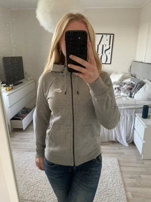 Grå zip hoodie från Nike - Snygg grå zip hoodie från Nike med vit logga på bröstet. Hoodien har dragkedja hela vägen, två fickor fram och justerbar huva med snören. Perfekt till chill eller streetwear-stil. Mjuk och skön bomullsblandning.