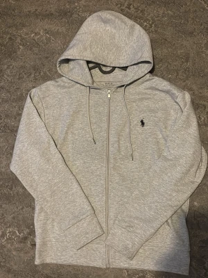 Grå zip hoodie från Polo Ralph Lauren - Snygg gråmelerad hoodie från Polo Ralph Lauren med dragkedja framtill och klassisk huva med dragsko. Broderad logga i svart på bröstet och två fickor framtill. Tillverkad i mjuk bomullsblandning som känns skön mot huden. Perfekt för en avslappnad och stilren look.