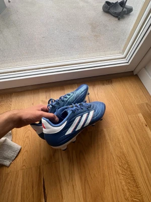 Blå Adidas Copa fotbollsskor - Säljer ett par blå Adidas Copa fotbollsskor med klassisk vit sula och vita ränder på sidan. Skorna har snörning och är tillverkade i syntetmaterial med en stilren design som passar perfekt för spel på gräsplan och konstgräs. Riktigt snygga och bekväma för dig som vill ta spelet till nästa nivå.