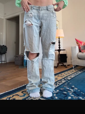 Ljusblå slitna baggy jeans - Säljer ett par ljusblå baggy jeans med stora slitningar och hål på både fram- och baksida. Jeansen har medelhög midja, raka ben och klassiska fem fickor. Perfekta för en avslappnad och trendig streetstyle-look.