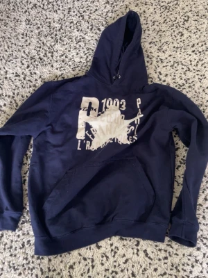 Zandro studio hoodie - Säljer en mörkblå hoodie med stor vit tryckt text och grafiskt motiv på bröstet. Hoodien har klassisk känguruficka framtill och justerbar huva med snören. Perfekt för dig som gillar streetwear och vill ha en avslappnad look. Nypris 1000kr, knappt använd!