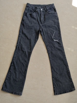 Svarta bootcut jeans med slitning - Snygga svarta bootcut jeans med hög midja och en cool slitning på ena benet. Klassisk femficksmodell och knappgylf. Perfekta för dig som gillar en avslappnad men trendig look. HELT OANVÄNDA jeans 