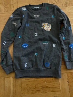 Grå sweatshirt från Kenzo med tryck - Cool grå sweatshirt från Kenzo med broderade och tryckta detaljer som tiger, ögon och bokstäver i blått, grönt och vitt. Tröjan har rund halsringning och ribbade muddar vid ärmslut och nederkant. Perfekt statement-plagg för dig som gillar streetstyle.