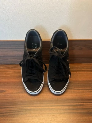 Svarta Polo Ralph Lauren sneakers - Säljer ett par svarta sneakers från Polo Ralph Lauren med klassisk låg profil och vit sula. Skorna har svart canvasmaterial, svarta skosnören och den ikoniska Polo-loggan broderad i guld på sidan. Perfekta för en clean och stilren look. De finns en grej där den är lite sliten och är på sista bilden