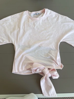 Ljusrosa t-shirt med knyt i sidan - Säljer en ljusrosa t-shirt från Gina Tricot med korta ärmar och en snygg knytdetalj i sidan. T-shirten har en klassisk rund halsringning och är gjord i mjuk bomull, perfekt för en avslappnad och trendig look.