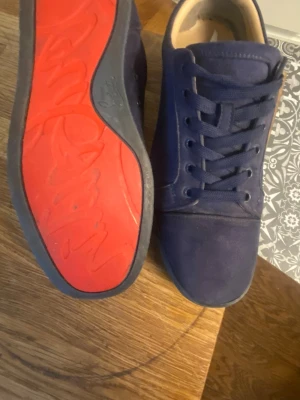 Mörkblå sneakers med röd sula - Snygga mörkblå sneakers med ovandel i mockaliknande material och klassisk snörning. Skorna har en unik röd yttersula med svart kant och diskret logga på sidan av sulan. Perfekta för dig som vill sticka ut med en cool detalj på fötterna. Har en litet bren hål där av priset 