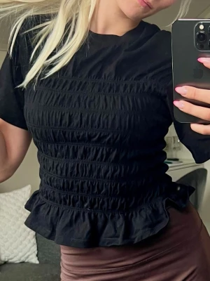 Svart topp med volangkant - Svart topp med korta ärmar och smockad midja som ger en figurnära passform. Toppen har en volangkant nedtill😊💕