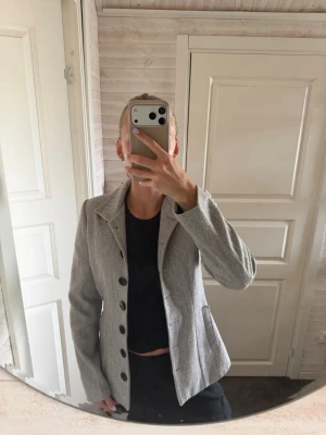 Ljusgrå kavaj med stora knappar - Snygg ljusgrå kavaj med markerad midja och stora svarta knappar framtill. Klassisk krage och två fickor på sidorna. Perfekt till både jeans och kostymbyxor för en stilren look. Materialet ser ut att vara en mjuk, vävd textil.