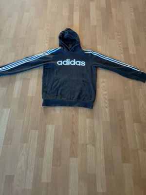 Svart Adidas hoodie med vita ränder - Säljer en svart hoodie från Adidas med klassiska vita ränder längs ärmarna och stor logga på bröstet. Hoodien har huva och magficka framtill. Perfekt för en sportig och avslappnad stil. Materialet känns mjukt och bekvämt.