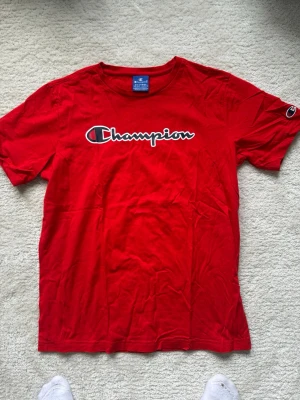 Röd Champion t-shirt i bomull - Snygg röd t-shirt från Champion med det klassiska logotrycket över bröstet och en liten logga på ärmen. Tillverkad i mjuk bomull och har en normal passform med korta ärmar. Perfekt för en avslappnad och sportig stil.