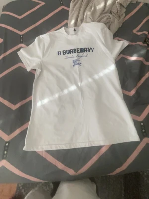 Vit Burberry t-shirt med tryck - Snygg vit t-shirt från Burberry med blått tryck på bröstet där det står 'Burberry London England'. Klassisk passform och rund hals. Perfekt till jeans eller shorts för en clean och stilren look.