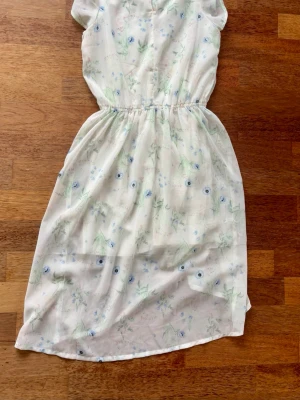 Vit blommig sommarklänning - Söt vit klänning med ljusblått och grönt blommönster. Klänningen har korta ärmar, resår i midjan och omlottkjol med lätt transparent material. Perfekt för varma dagar och har en luftig, flowy känsla.