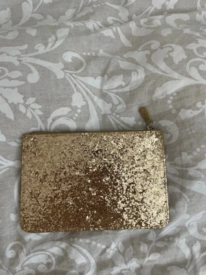 Glittrig guld plånbok  - Säljer en glittrig plånbok i guld med dragkedja. Plånboken har en rektangulär form och är täckt av glittrigt material som verkligen sticker ut. 