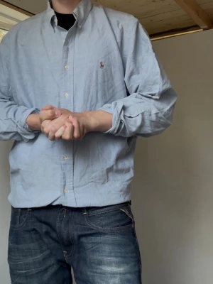 Ralph Lauren | Skjorta | L - Klassisk ljusblå skjorta från Ralph Lauren med button-down krage och broderad logga på bröstet. Perfekt för dompa & Grisch. Skjortan har långa ärmar, normal passform och är tillverkad i mjuk bomull. Perfekt för en clean och stilren look. Mått: 75 Längd, 63 Bredd