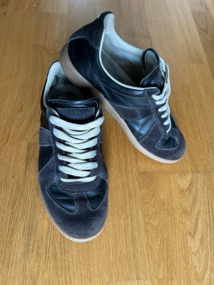 Grå sneakers med mockadetaljer - Snygga sneakers i grå mocka och svart läder med vita skosnören. Klassisk design med låg profil och gummisula. Perfekta för dig som gillar stilrena och tidlösa skor. Passar till det mesta och ger en clean look.