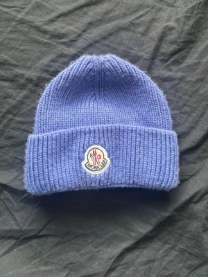 Blå ribbstickad mössa från Moncler - Säljer en blå ribbstickad mössa från Moncler med uppvikt kant och klassisk Moncler-logga framtill. Mössan är gjord i mjukt material och har en enkel, stilren design som passar perfekt till kalla dagar.