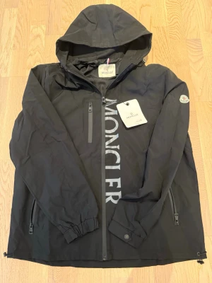  Moncler - Snygg svart vindjacka från Moncler med stor logga framtill och klassisk Moncler-patch på ärmen. Jackan har huva, dragkedja, två sidofickor med dragkedja och elastiska muddar. Materialet är lätt och vindtätt, perfekt för blåsiga dagar.