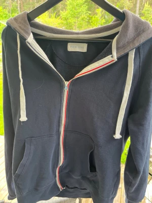 Mörkblå zip-hoodie från Peer One - Snygg mörkblå hoodie från Peer One med dragkedja och vita snören. Insidan av huvan är grå och har ett mjukt, strukturerat tyg. Två stora fickor framtill och en rödvit detalj längs dragkedjan. Perfekt för chill dagar eller när du vill ha en avslappnad stil. Kan gå ner lite i pris
