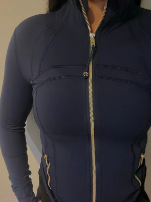 Mörkblå ziptröja från Lululemon - Snygg mörkblå ziptröja från Lululemon med guldfärgade dragkedjor och detaljer. Tröjan har figurnära passform, hög krage och två fickor med dragkedja. Perfekt för träning eller som ett sportigt lager till vardags.