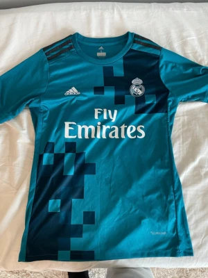Real Madrid 17/18 T-shirt - (Pris kan diskuteras) Säljer en blå Real Madrid matchtröja från Adidas med Ronaldo och nummer 7 på ryggen. Tröjan har korta ärmar, klassiska Adidas-ränder på axlarna och Fly Emirates-tryck på bröstet. Tillverkad i ett lätt och funktionellt material som passar perfekt till fotbollsträning.