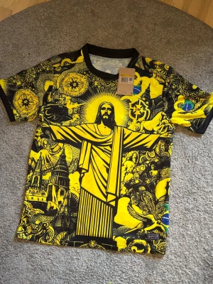 Gul Brasilien t-shirt med tryck - Cool gul t-shirt från Nike med Brasilien-tema och stort grafiskt tryck av Kristusstatyn, Rio och CBF-loggan. Svarta detaljer vid krage och ärmslut. Perfekt för dig som gillar unika och färgstarka plagg med fotbollsvibbar.