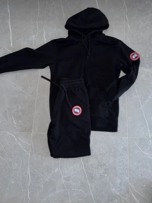 Svart loungewear-set från Canada Goose - Snyggt svart loungewear-set från Canada Goose med matchande hoodie och byxor. Båda plaggen har den klassiska röda Canada Goose-loggan. Hoodien har dragsko och magficka, byxorna har resår i midjan och dragsko för skön passform. Perfekt för chill dagar hemma.