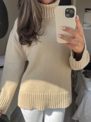 Beige polotröja med breda muddar - Säljer en beige stickad polotröja med bred ribbstickad krage, ärmslut och nederkant. Tröjan har en avslappnad passform och långa ärmar. Perfekt för kalla dagar och enkel att styla med jeans eller kjol.
