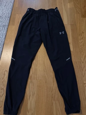 Svarta träningsbyxor Under Armour - Säljer ett par svarta träningsbyxor från Under Armour med resår i midjan och reflexdetaljer på benen. Byxorna har smal passform, diskret logga på låret och är tillverkade i ett lätt och stretchigt material som passar perfekt för löpning eller gym.