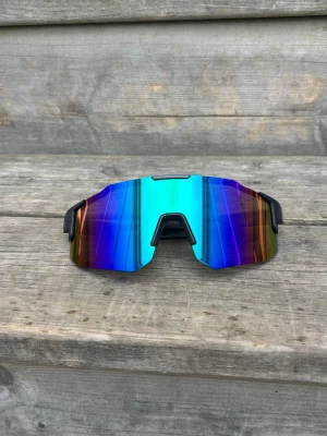 Sportiga spegelglas solglasögon - Säljer ett par sportiga solglasögon med stora, spegelblå linser och svarta bågar. Glasen har en cool regnbågseffekt i blått, lila och grönt. Perfekta för dig som vill sticka ut och få en futuristisk vibe på sommaren.