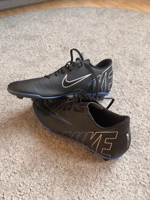 Nike Mercurial svarta fotbollsskor - Säljer ett par svarta Nike Mercurial fotbollsskor med stora vita loggor på sidorna och blå detaljer på sulan. Skorna har snörning och är tillverkade i ett lätt syntetmaterial som ger bra bollkontroll och grepp på planen. Perfekta för dig som vill ha stilrena och snabba skor.
