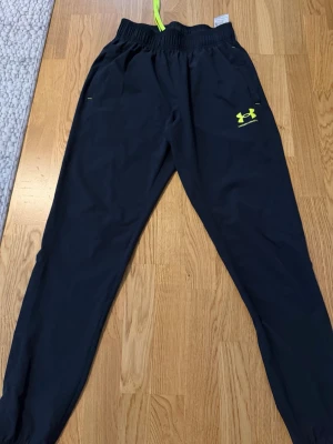 Svarta mjukisbyxor från Under Armour - Säljer ett par svarta mjukisbyxor från Under Armour med neongula detaljer och logga på benet. Byxorna har resår i midjan med justerbar dragsko och smal passform. Perfekta för chill eller träning, tillverkade i ett lätt och bekvämt material.