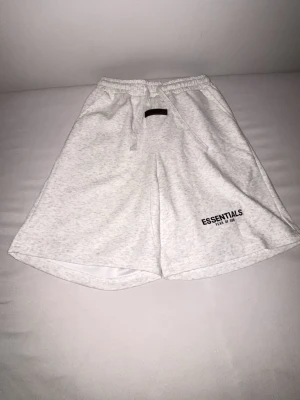 Vita Essentials shorts med snörning - Säljer ett par ljusa, vita shorts från Essentials med tryckt logga på benet. De har elastisk midja med snörning och en bakficka. Sköna och avslappnade, perfekta för sommaren. Materialet känns mjukt och är bomull eller bomullsblandning. 