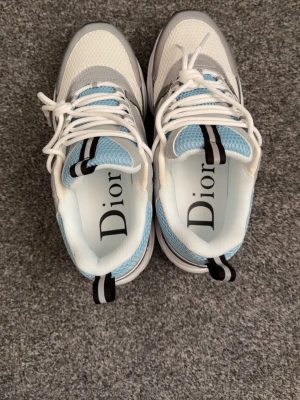 Vita CD skor - Snygga chunky sneakers i vitt, ljusblått och grått. Ovandelen är i mesh och läder med coola detaljer och svart/vit dragflik bak. Tjock sula för extra streetstyle-vibe och sportig look. Perfekta för dig som vill sticka ut med exklusiva sneakers.