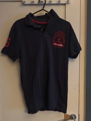 Mörkblå Peak Performance piké - Mörkblå pikétröja från Peak Performance med röd broderad logga och text på bröstet samt siffra på ärmen. Klassisk krage och korta ärmar. Perfekt för en sportig och avslappnad stil.