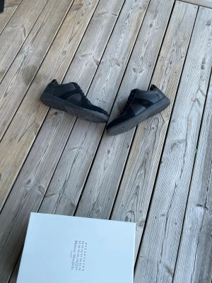 Svarta sneakers från Maison Margiela - Snygga helsvarta sneakers från Maison Margiela med klassisk låg silhuett. Skorna har en mix av mocka och läder på ovandelen samt en robust gummisula med tydligt mönster. Perfekta för dig som gillar minimalistisk och stilren design.