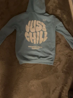 Blå hoodie med tryck 'Just Chill' - Ljusblå hoodie med stor vit text 'JUST CHILL' och 'Escaping from reality' på ryggen samt mindre tryck framtill. Klassisk känguruficka och huva. Perfekt för en avslappnad stil och sköna dagar. Mjuk bomullsblandning och ribbade muddar.