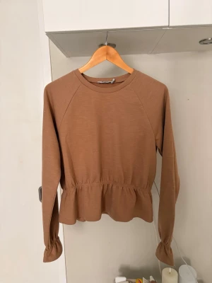 Brun volangtopp från NA-KD - Supersöt brun topp från NA-KD med volang nertill och långa ärmar med knyt vid handlederna. Perfekt för dig som gillar en feminin och trendig stil. Tillverkad i mjukt material som känns skönt mot huden. 🥰🥰