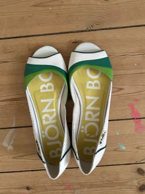 Ballerinaskor Björn Borg - Snygga ballerinaskor från Björn Borg i vitt med gröna och turkosa detaljer. Skorna har öppen tå, perforerade sidor och en gul innersula med stort logotryck. Tillverkade i konstläder med metallmärke på sidan. Perfekt för dig som gillar sportig och färgglad stil.