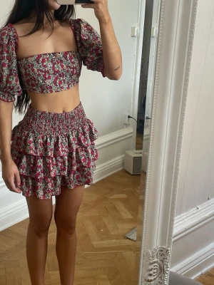 Blommigt tvådelat set med volangkjol - Säljer ett superfint tvådelat set med blommönster i rosa, vitt och grönt. Toppen har puffärmar och rak urringning, kjolen är kort med smock i midjan och dubbla volanger. Perfekt för dig som gillar trendiga och feminina plagg!