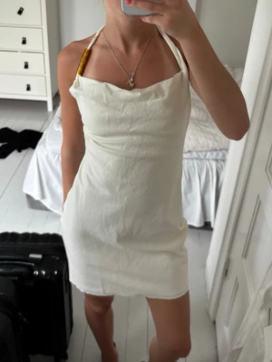 Vit halterneck kortklänning - Säljer en stilren vit halterneck klänning med kort längd och öppen rygg. Klänningen har en enkel, figurnära passform och tunna axelband som knyts i nacken. Perfekt för varma dagar och sommarens alla tillfällen.
