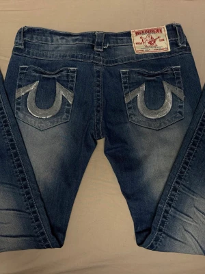 True Religion skinny jeans med paljettdetaljer - Säljer ett par blå jeans från True Religion med ikoniska bakfickor dekorerade med silvriga paljetter. Jeansen har en klassisk femficksmodell och en snygg tvätt med slitningar. Perfekta för dig som vill sticka ut med coola detaljer. Storlek 30, skinny modell.