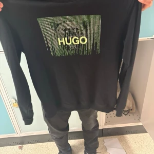 Svart sweatshirt från HUGO BOSS - Svart sweatshirt från HUGO med ett stort grönt och gult grafiskt tryck och logga på bröstet. Tröjan har rund halsringning och långa ärmar. Perfekt för en trendig och avslappnad stil. 