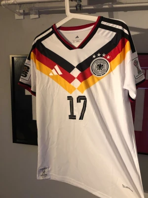 Tysklands matchtröja Adidas Wirtz 17 - Snygg vit matchtröja från Adidas för Tysklands landslag med nummer 17 och namnet Wirtz på ryggen. Tröjan har svarta, röda och gula detaljer, V-ringning och officiella märken på ärmarna. Tillverkad i lätt och ventilerande material, perfekt för fotboll.