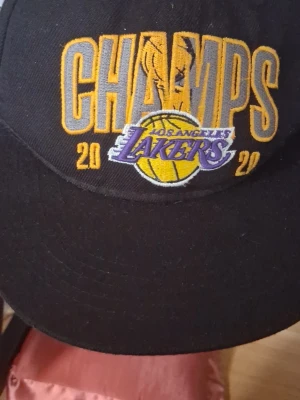 Svart Lakers Champs keps New Era - Säljer en svart keps från New Era med broderad text 'CHAMPS' och Los Angeles Lakers-logga framtill. Kepsen har gula och lila detaljer samt årtalet 2020 broderat. Perfekt för dig som älskar basket och vill representera Lakers.