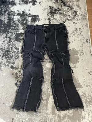 Svarta patchwork jeans med råa kanter - Unika svarta jeans med patchwork-design och råa, fransiga kanter över hela byxan. Modellen har raka ben och flera synliga sömmar som ger en edgy och trendig look. Perfekta för dig som vill sticka ut med din stil. Price when brand new 150-160€